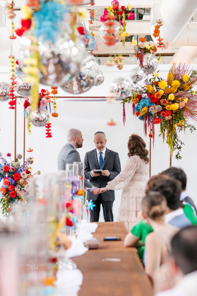 Bride and groom under a colorful flower-adorned wedding chuppah