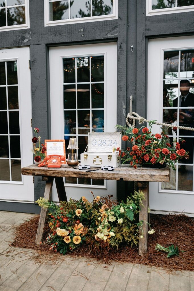 A hand-hewn wooden table containing many sweet wedding details: A case with the date 9/28/24 engraved in it, an old-fashioned orange rotary telephone, disposable cameras, and more. Three rustic, autumnal flower arrangements adorn the setup.