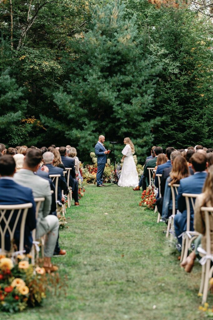 Rex and Jess at their autumnal flower-adorned altar before a majestic wooded backdrop, with their loved ones looking on.