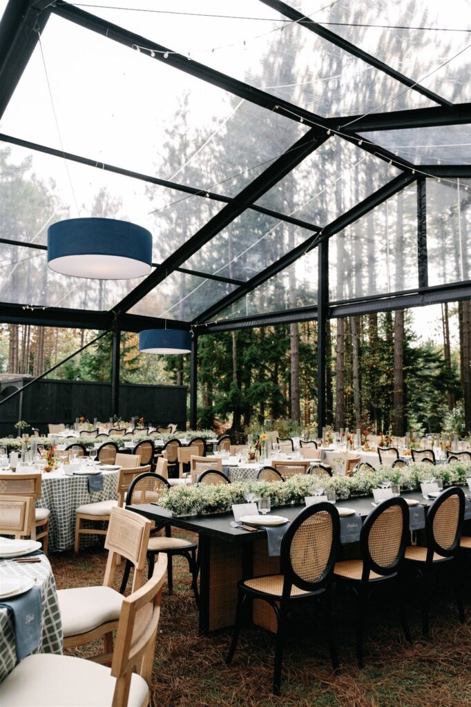 A black-framed A-frame tent with transparent walls looking on to a stretch of woods. Inside the tent are several dining tables set with floral centerpieces.