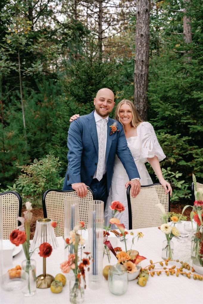 Rex in a blue suit and Jessica in a white gown, standing together and smiling between wooded background and a table adorned with autumnal bud vase arrangements and scattered fruits.