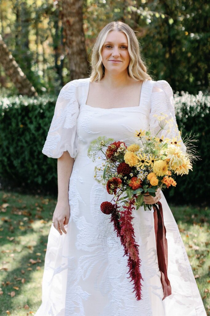 Jessica in a white gown with ethereal bell half-sleeves. She holds an autumnal bouquet tied together with a bordeaux silk ribbon.