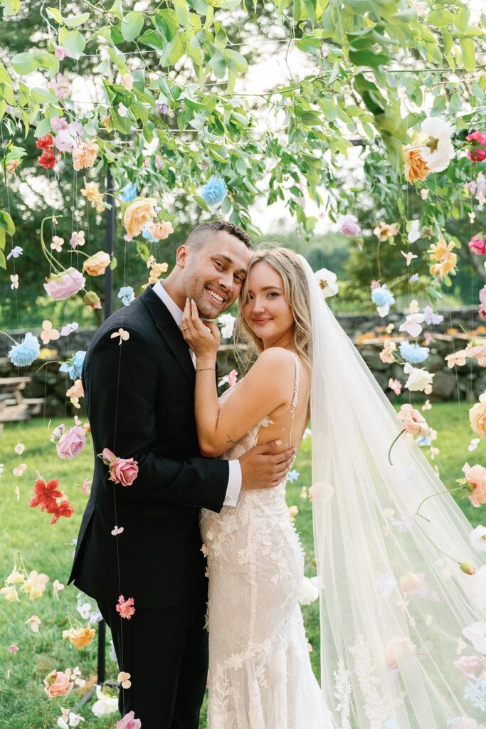 A bride and groom embracing and smiling at the camera amidst hundreds of hanging flowers.