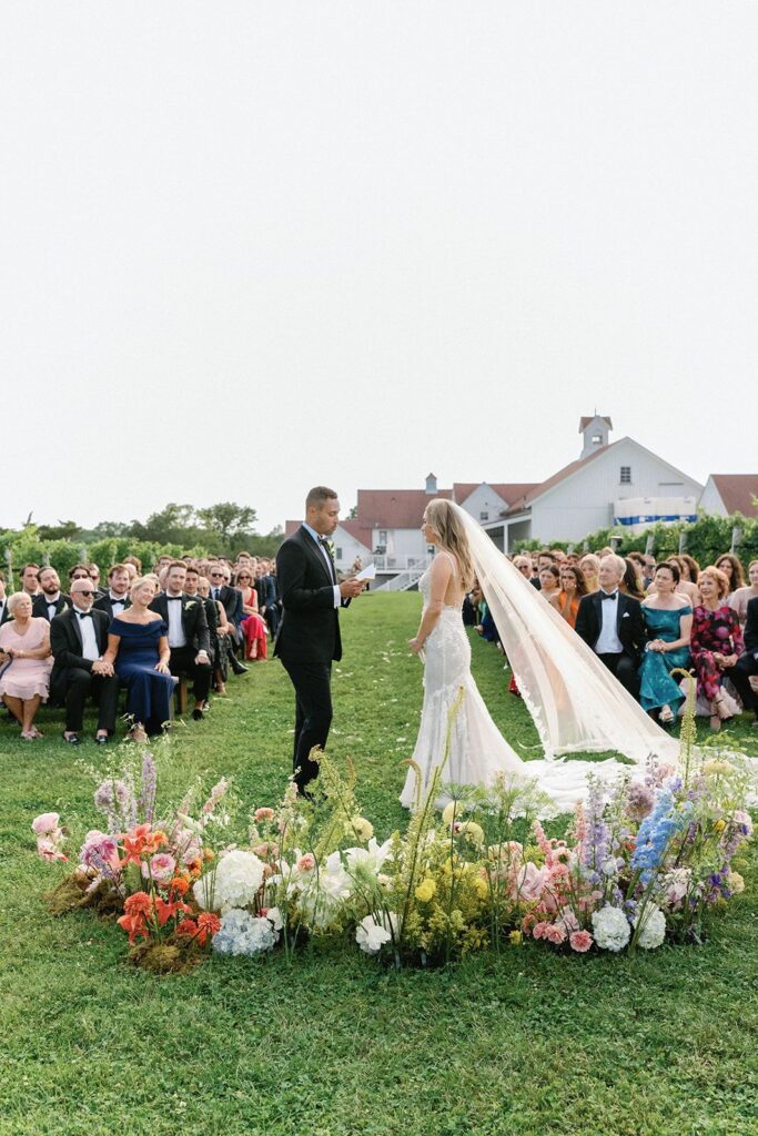 A bride and groom standing before an eclectic, color-blocked rainbow ground arch of flowers with all their seating loved ones looking on.