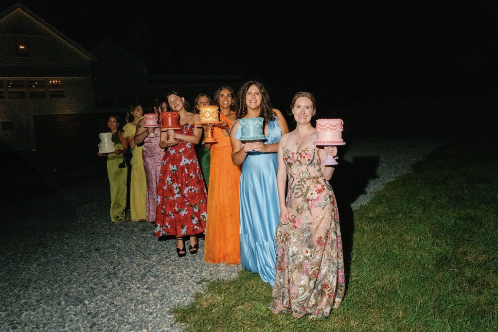 A line of bridesmaids in a rainbow of different-colored dresses, each holding a cake frosted to match her dress