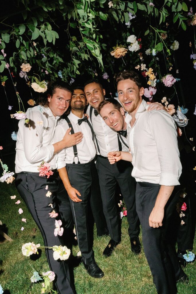 A group of tuxedo'd gentlemen smiling beneath a cascading canopy of flowers