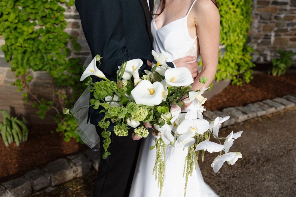 A bride and groom photo zoomed in on the bride's white bouquet. It contains arum lilies, orchids, daffodils, bells of Ireland, allium stems, and amaranthus strands.