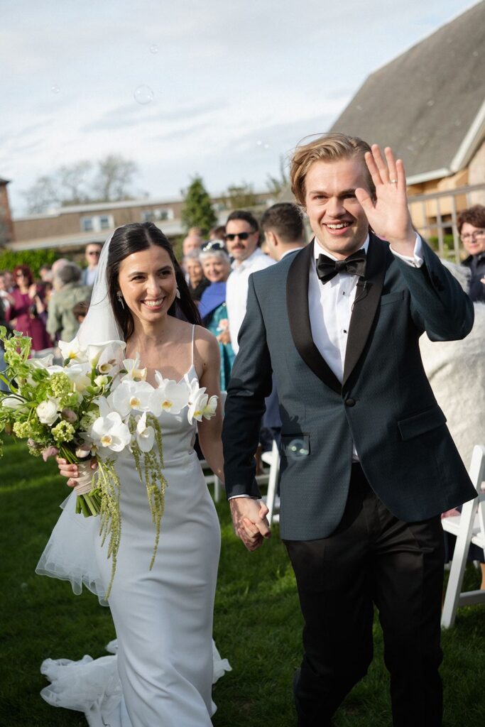 A bride and groom waving to their guests as they walk down the aisle of their outdoor wedding. The bride holds a white bouquet with orchids, arum lilies, and lots of greenery.