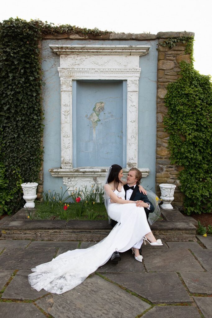 A bride and groom sitting before a pale blue fountain-like architectural moment built into a stone wall.