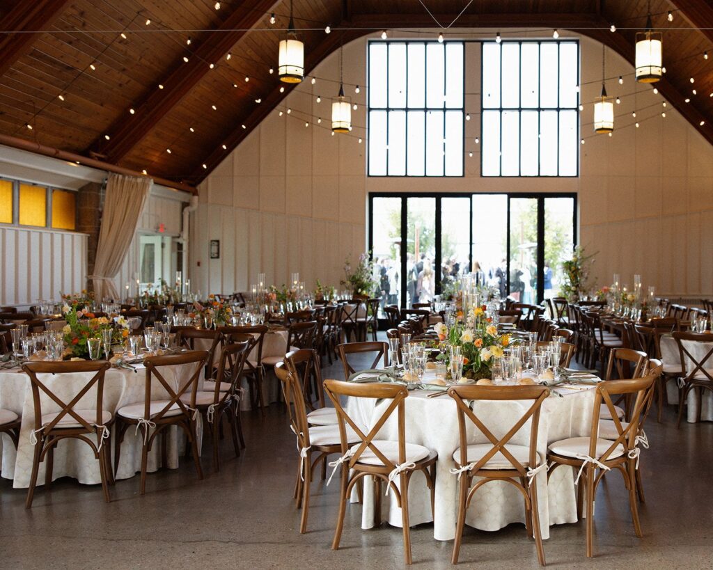 A giant barn-like dining room with high wooden ceilings, big picture windows, and tiny fairy lights strung across the space. Beneath the lights are several set tables with elaborate floral centerpieces.