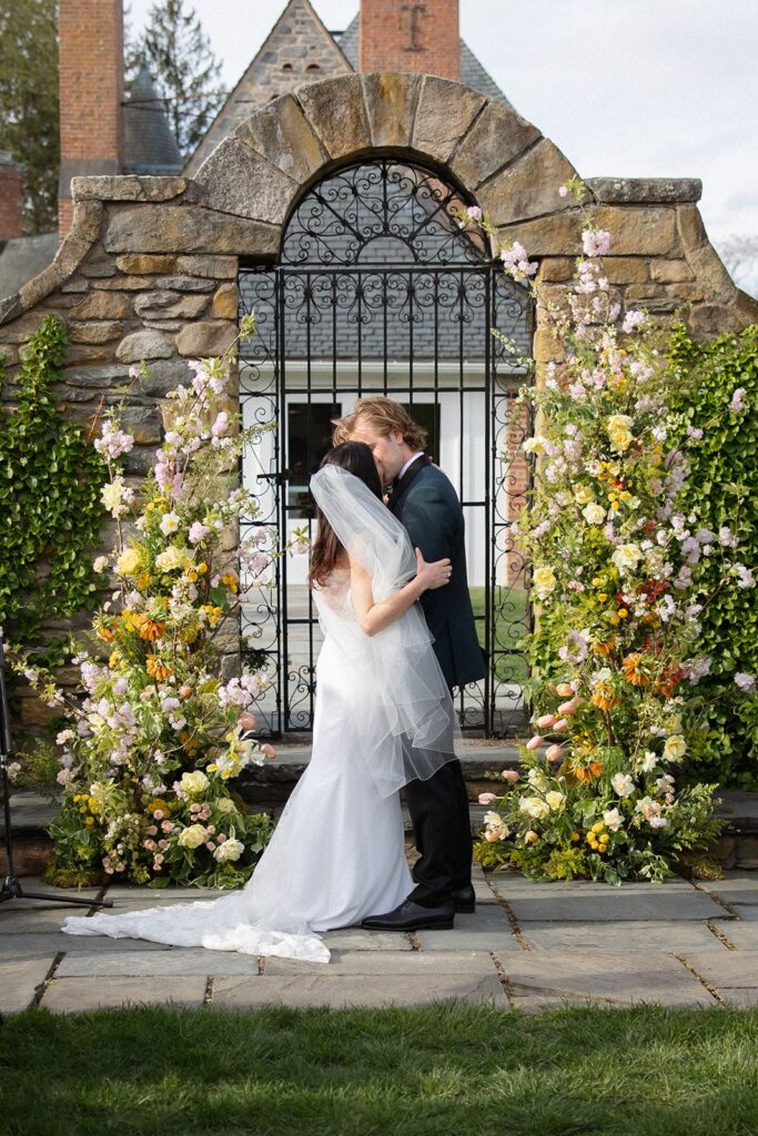 A bride and groom share their first kiss at their outdoor wedding. Behind them is elaborate castle-like stonework and a deconstructed arch of spring flowers.