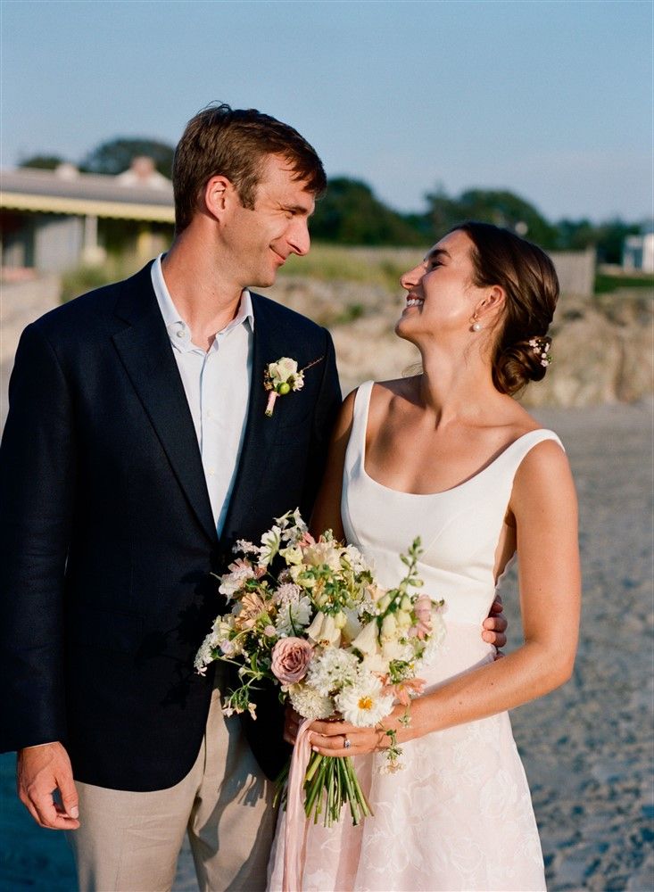 A bride and grooom on the beach. The groom wears beige pants, a blue shirt, and a navy jacket with a pale yellow and peach boutonniere. The bride wears a gown with a white top and peach skirt with appliqué flowers, and holds a pastel-colored bouquet.