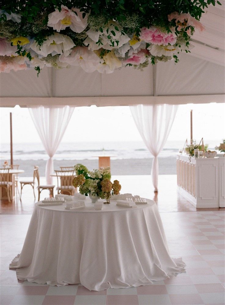 A wedding reception in a solid-floor tent by the beach. In view is a white-clothed table with a dynamic hydrangea centerpiece and a floral overhang with giant paper flowers.