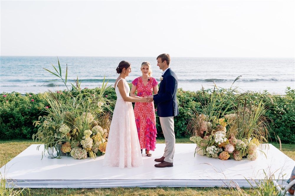 A bride and groom being married by a woman in fuchsia at an oceanside ceremony. On either side of the bride and groom are ceremonial floral installs crafted with hydrangeas and long grasses.