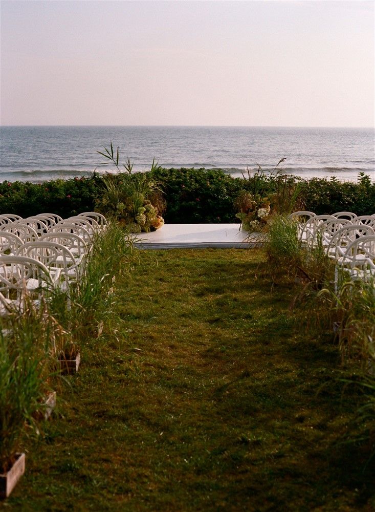 A grassy oceanside wedding aisle lined with tall grasses toward an altar decorated with more tall grasses and hydrangeas.